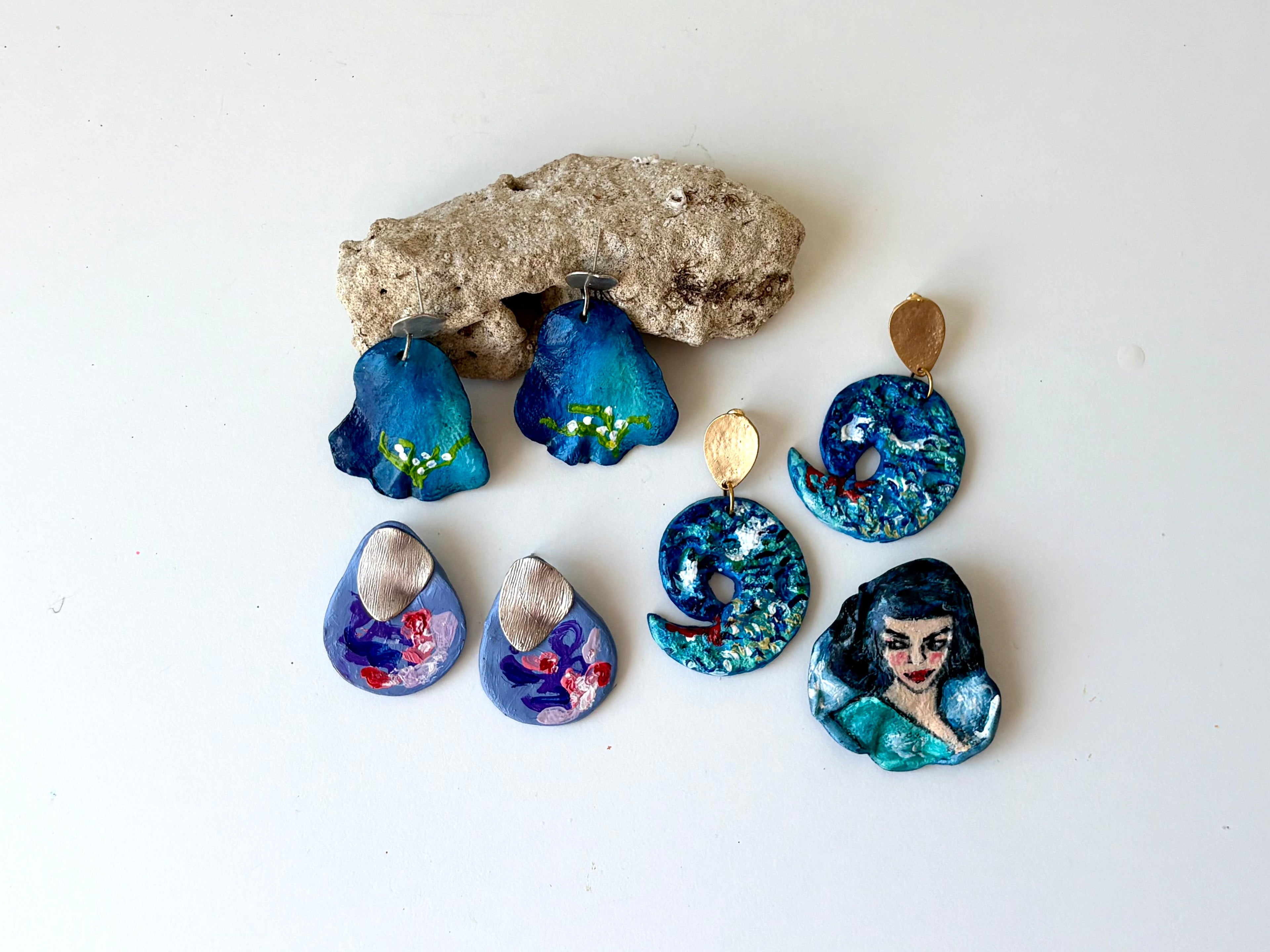 A set of handmade earrings displayed on a plain white background, featuring blue and purple floral, mermaid, and abstract designs with gold-tone pendants.